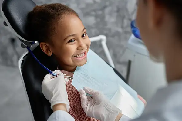 A smiling, excited young beautiful Black girl sits in the dental chair, eagerly awaiting her dental exam while her dentist places a bib on her.
