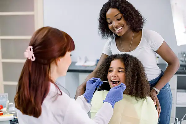 A pediatric dentist examines a calm girl's mouth while her mother watches during a routine dental checkup.