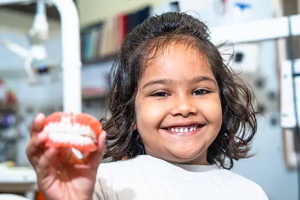 A happy young girl smiling and holding up a set of dentures, revealing her comfort with dentistry for kids.