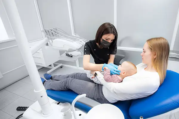 A young woman supports her baby daughter in a dentist chair while a dentist inspects her child's mouth.