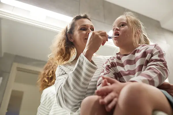 A mother helping her special needs daughter brush her teeth in a bright bathroom.